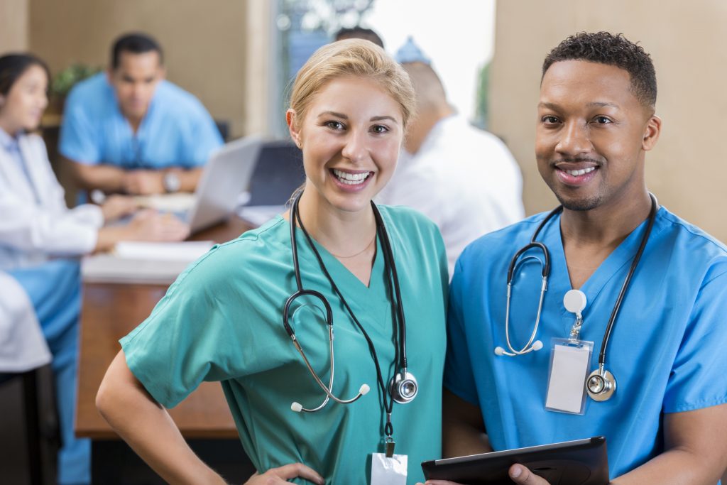 Young Caucasian female and male mid adult African American nurses smile before hospital staff meeting. The woman is wearing green scrubs and the man is wearing blue scrubs. They are both wearing stethoscopes. People are talking in the background.